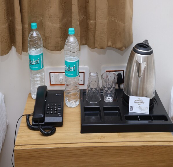 Habitación de hotel con un escritorio que contiene un teléfono, dos botellas de agua Bisleri, dos vasos de cristal, una tetera de acero inoxidable y una bandeja con tetera y taza.
