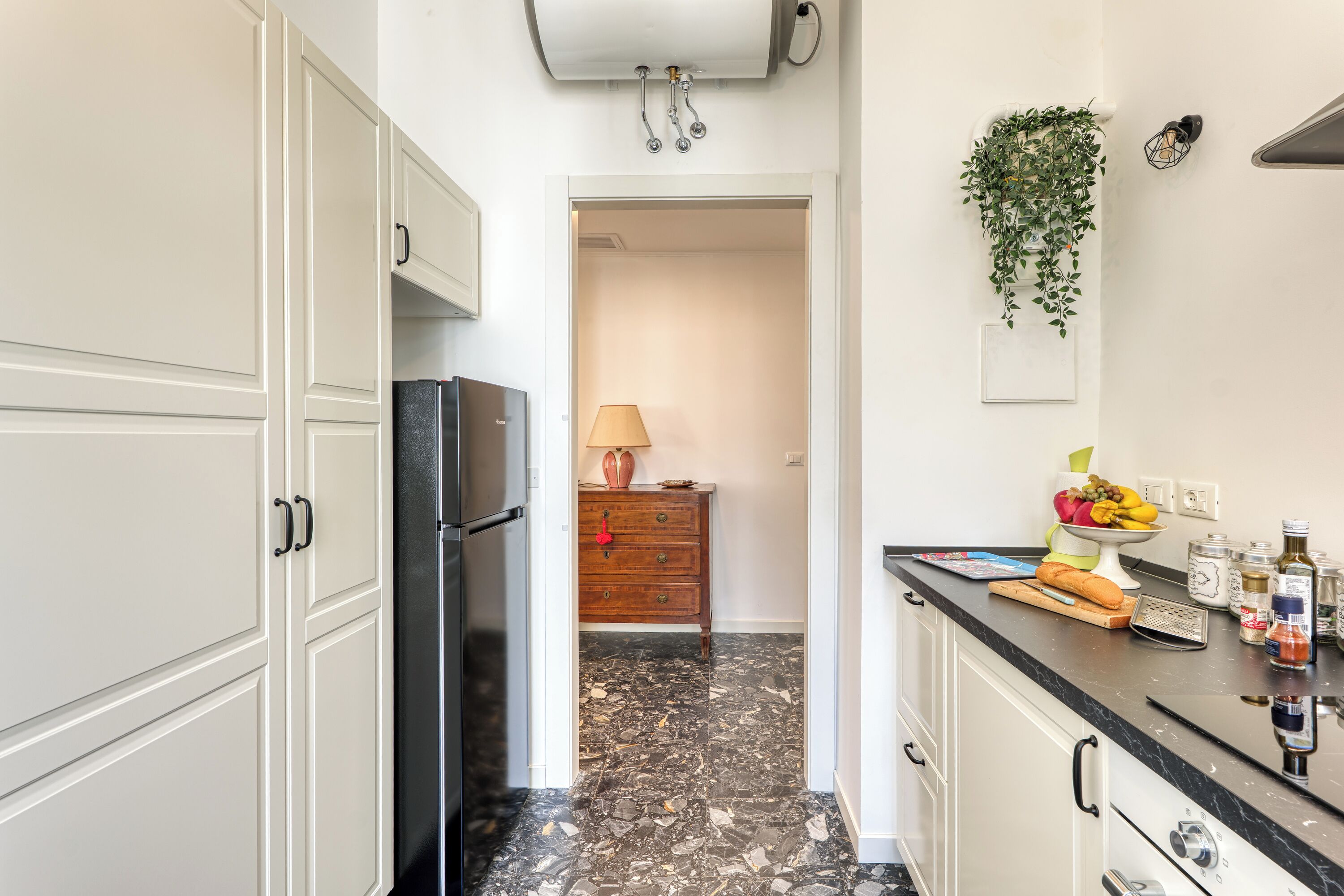 A kitchen with white cabinets, a black refrigerator, a wooden dresser, and a countertop with various items.