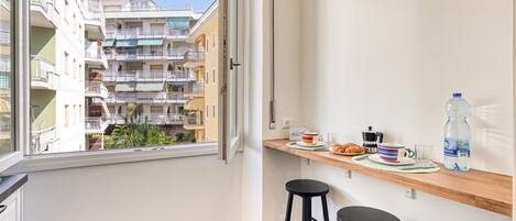 Un balcon donnant sur un immeuble à plusieurs étages, une étagère en bois avec des articles pour le petit-déjeuner et un tabouret noir.