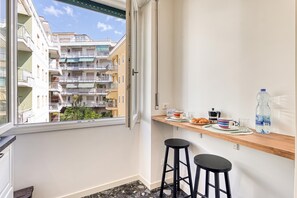 A balcony with a view of a multi-story building, a wooden shelf with breakfast items, and a black stool.