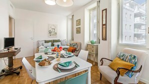 Una sala de estar moderna con comedor, sofá, televisor y ventana con vista a edificios.
