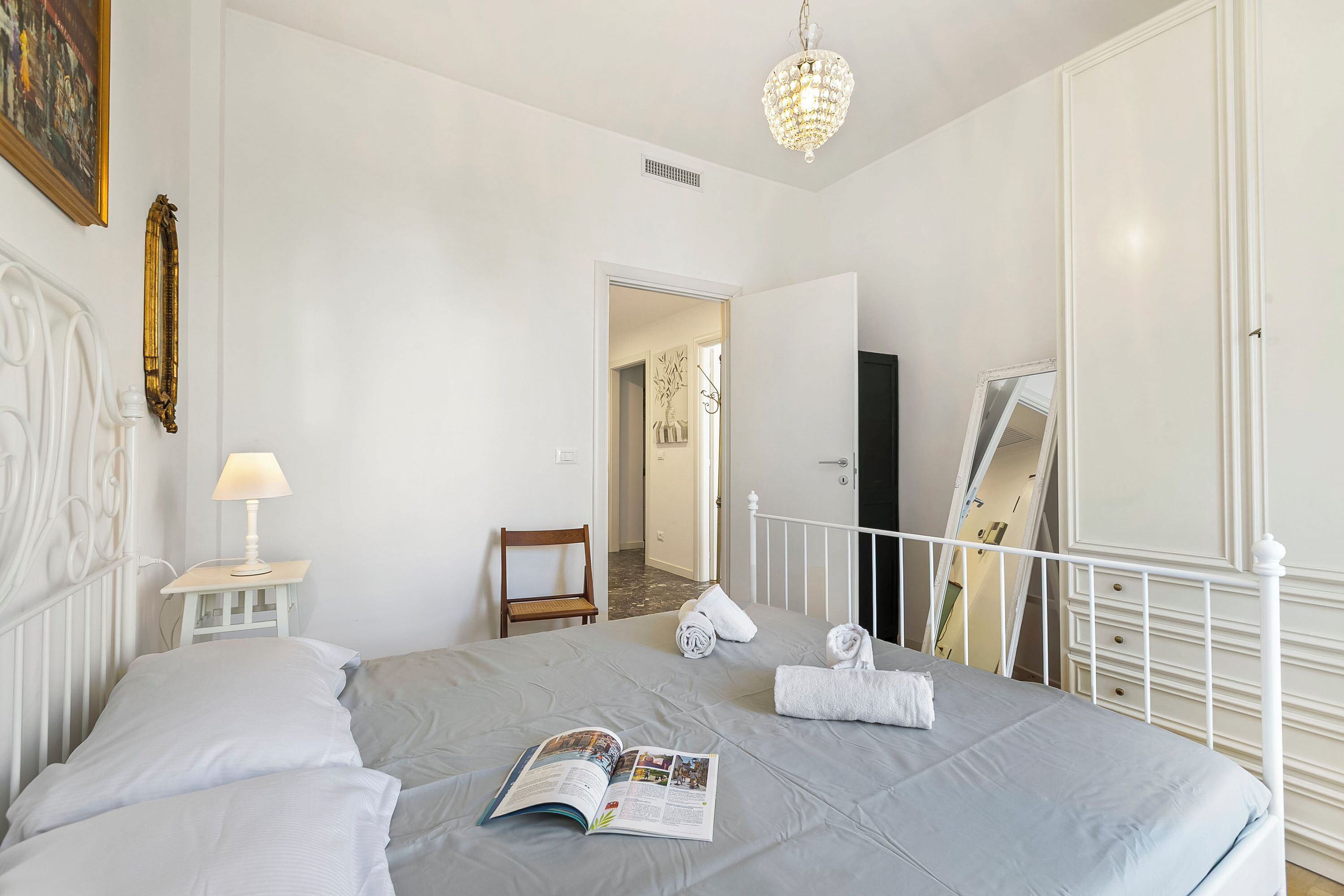 A bedroom with a bed, a bedside lamp, a book, a towel, and a mirror.