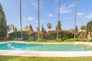 Una piscina amb superfície d'aigua transparent i blava, envoltada per gespa ben cuidada, palmeres i arbustos florits.