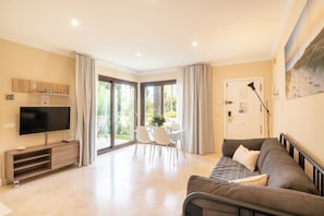 A modern living room with a flat-screen TV, a sofa, a dining table with chairs, and a large window with curtains.