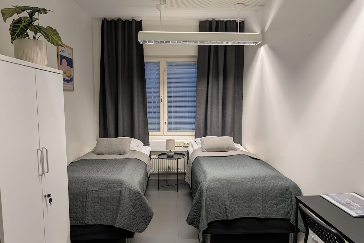 A small, modern bedroom with two single beds, a desk, a chair, and a window with curtains.