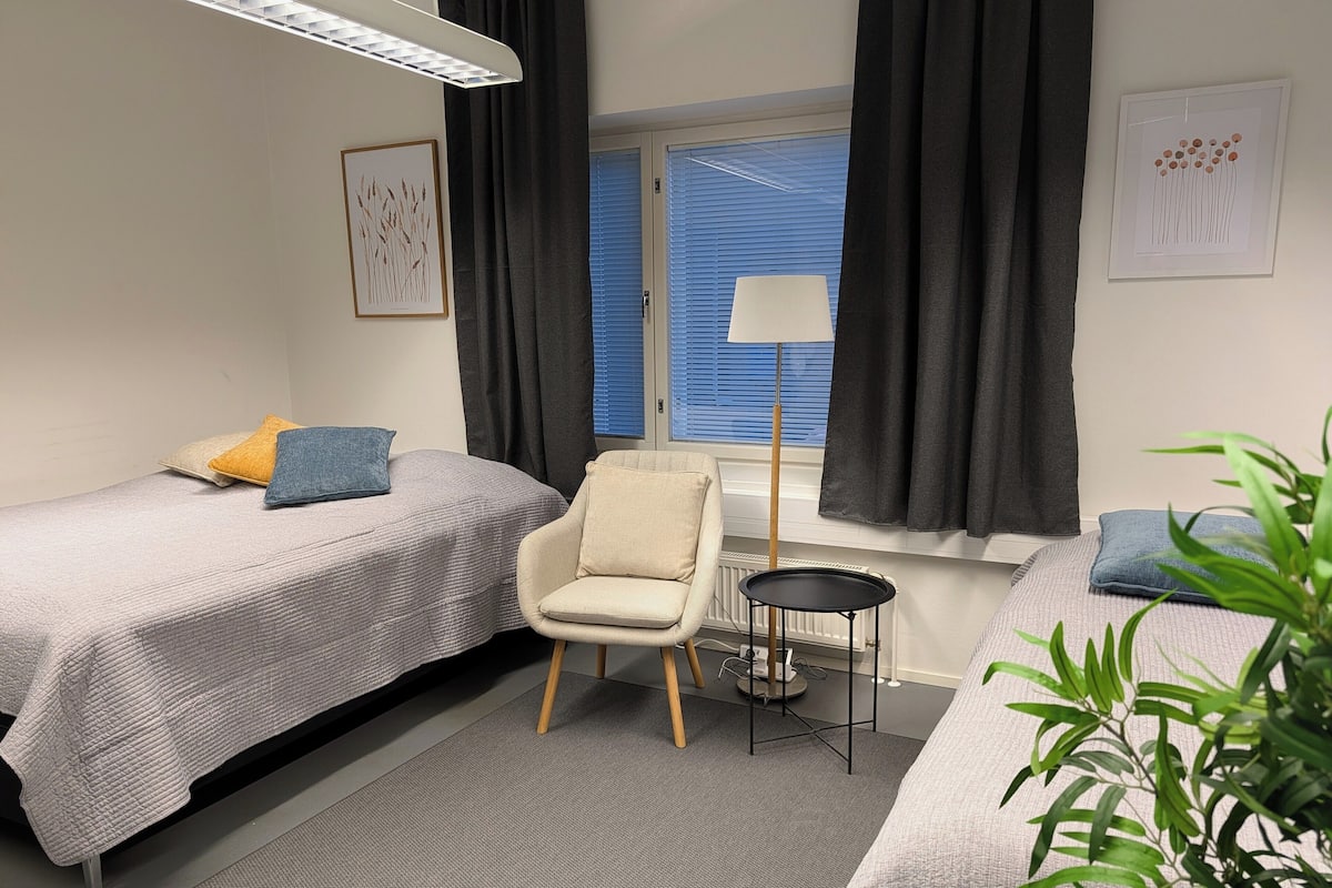 A modern bedroom with two beds, a chair, a floor lamp, and a window with curtains.
