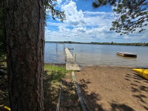 En brygga vid sjön med en träpir som sträcker sig ut i det stilla vattnet, omgiven av träd och en klar himmel.