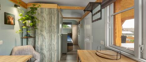 A narrow hallway with a wooden ceiling, a dining area with a table and chair, a bed in the background, and a window with a view.