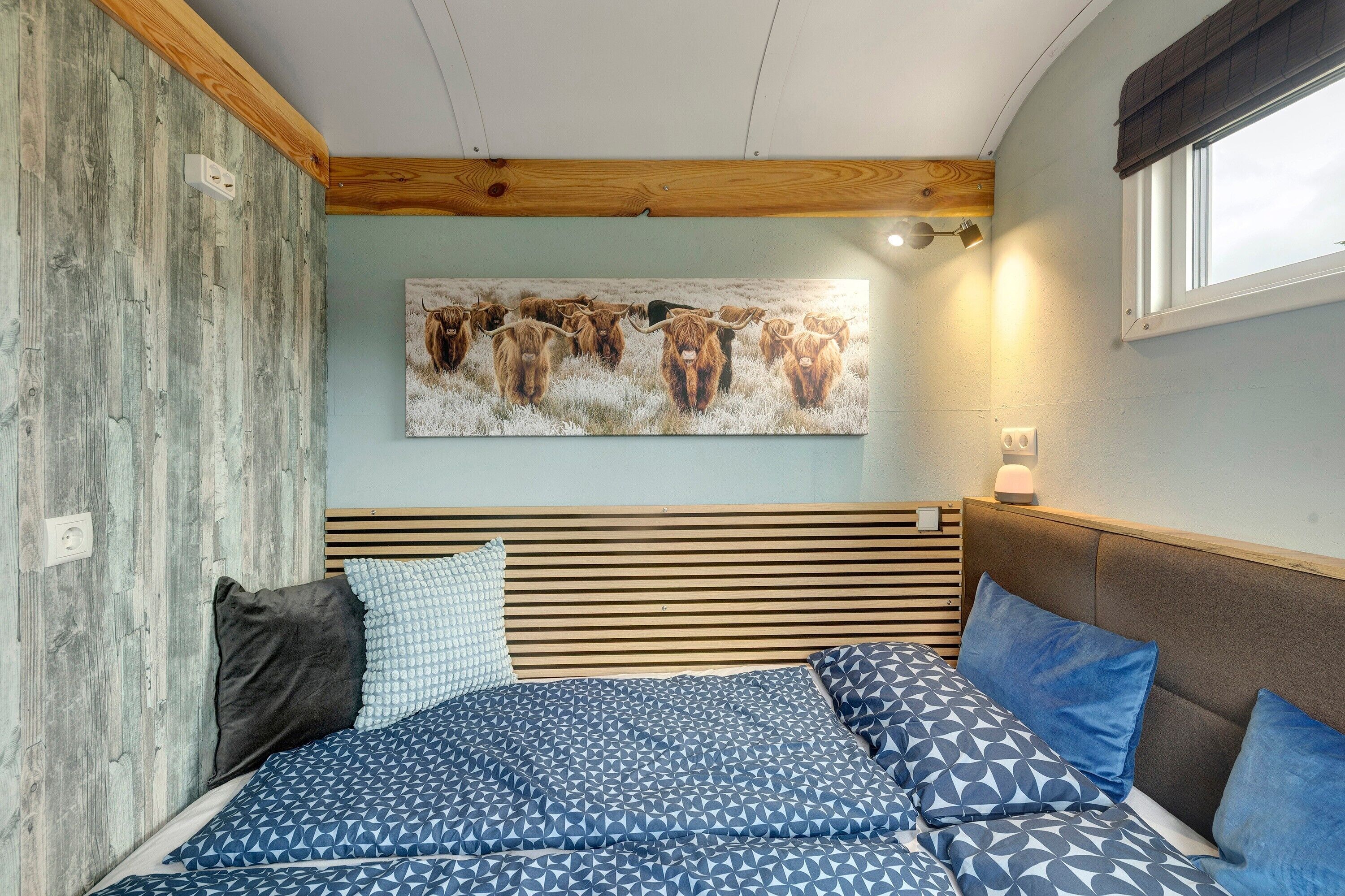 Una camera da letto con testiera in legno a listelli, un letto con biancheria a fantasia, un quadro appeso al muro raffigurante bovini e una finestra con vista.