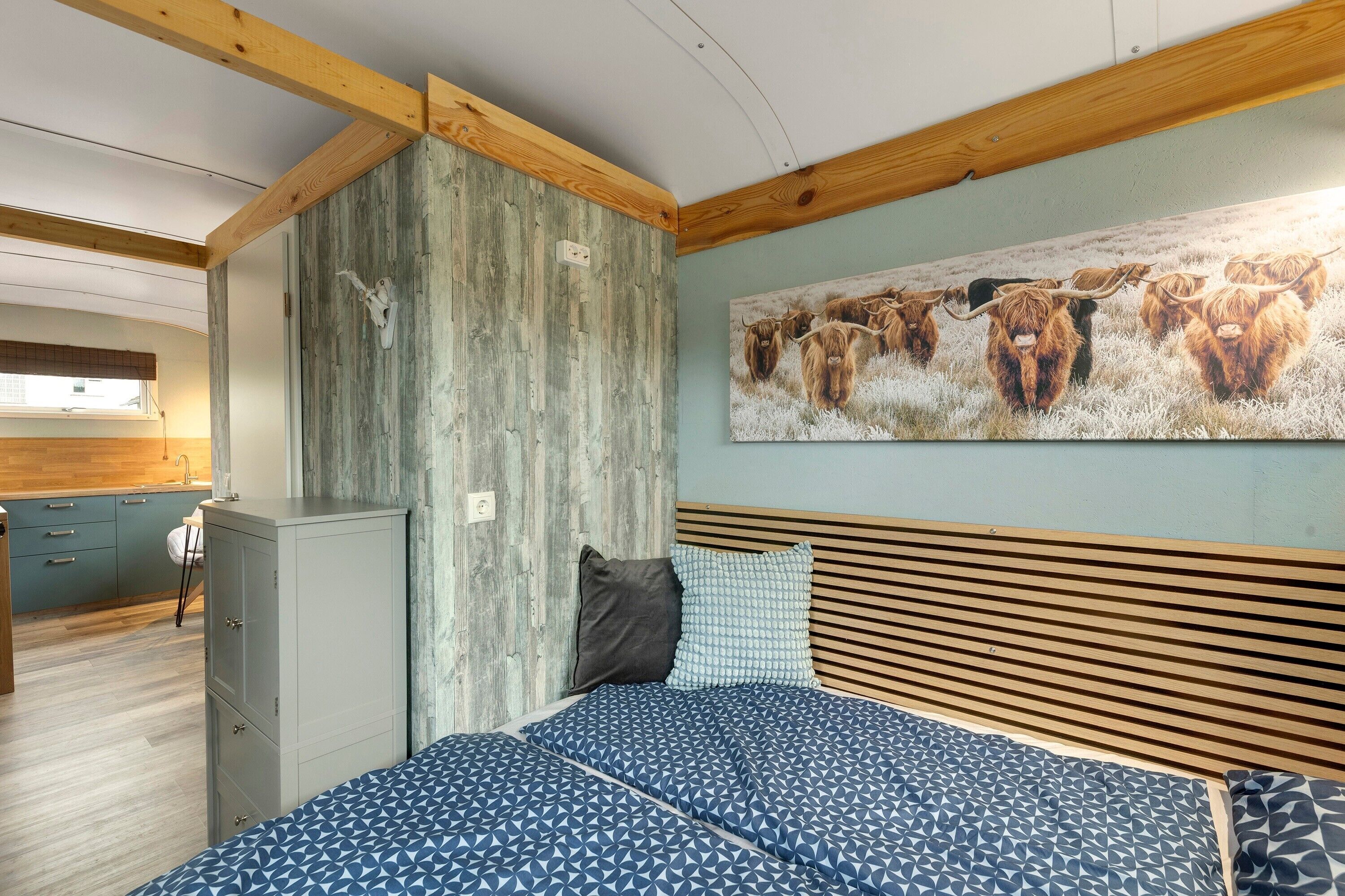 Une chambre à coucher avec un lit, une tête de lit en bois, un tableau représentant des yaks accroché au mur, une armoire et, au fond, une petite cuisine.