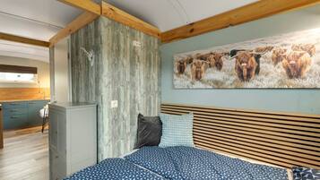 A bedroom with a bed, wooden headboard, a wall-mounted painting of yaks, a wardrobe, and a kitchenette in the background.