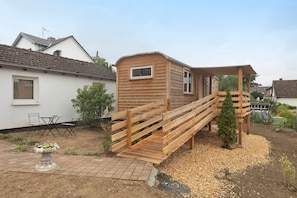 Een houten terras met een trap naar een klein houten gebouw, omgeven door een tuin met een bloempot en een tafel met stoelen.