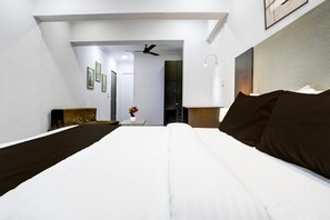 Ein Hotelzimmer mit einem großen Bett, einem Deckenventilator, einem kleinen Tisch mit Blumenvase und Blick auf den Flur.