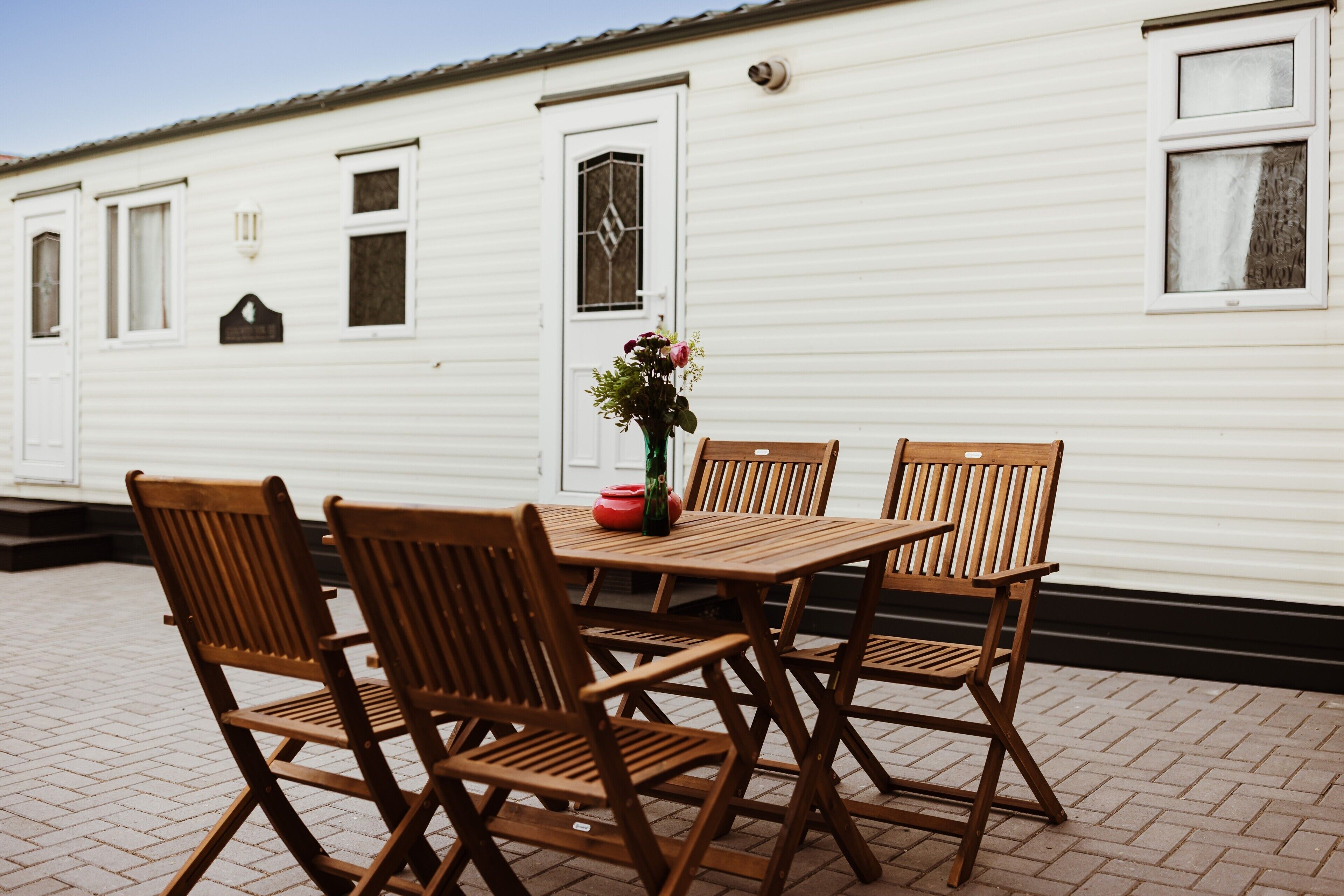 Une maison mobile blanche avec une terrasse en briques, une table en bois avec des chaises et un vase rempli de fleurs.