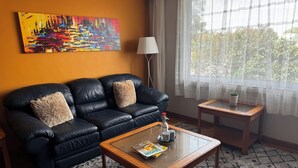 Un salon avec un canapé en cuir noir, deux coussins moelleux, une table basse avec des magazines, une lampe et une fenêtre ornée de rideaux légers.