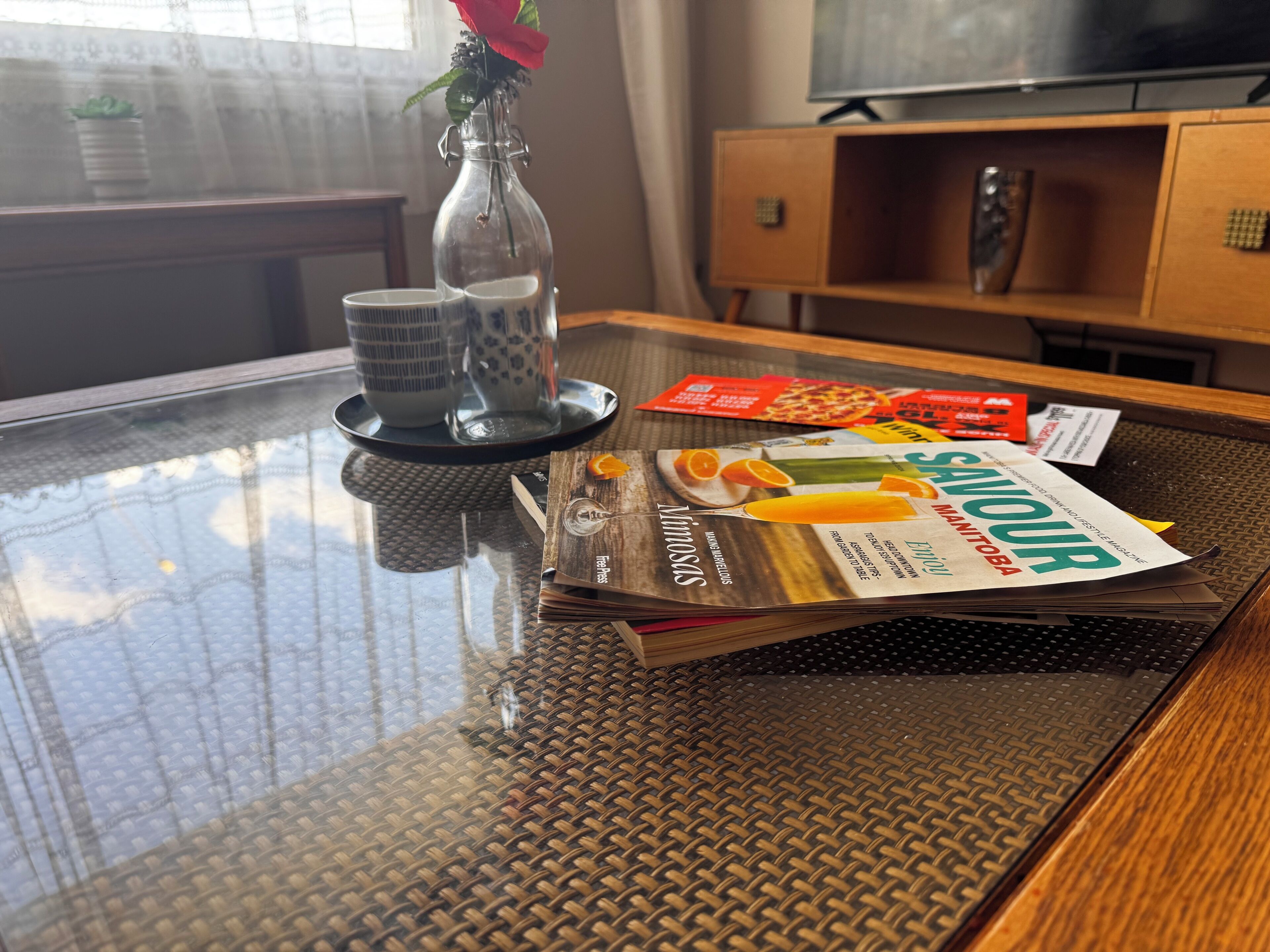 A glass-top dining table with a magazine, a vase with a red flower, and a tray with a pizza box and a magazine.
