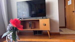 Une pièce avec un meuble TV en bois, un téléviseur à écran plat, un vase en verre contenant une fleur rouge et un magazine posé sur le sol.
