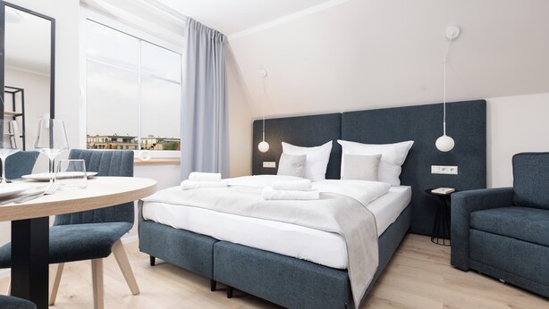 Habitación de hotel moderna con una cama grande, una mesa de comedor redonda y vistas a la ciudad a través de amplios ventanales.