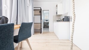 Una habitación de hotel moderna con una mesa de comedor de madera, sillas tapizadas en gris, una cocina con lavabo y encimera, y vistas a través de una puerta abierta.
