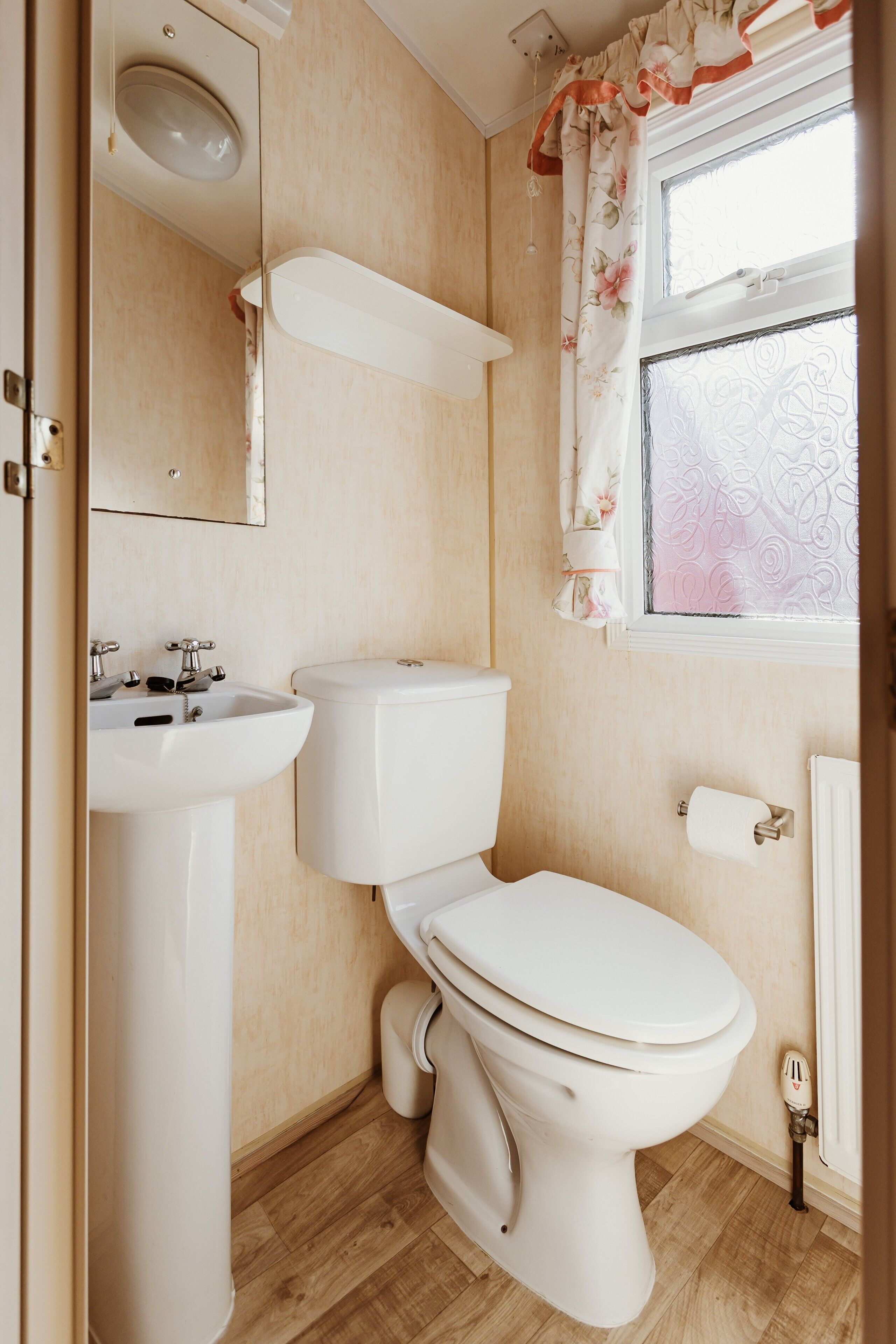 A bathroom with a toilet, sink, mirror, and window with floral curtains.