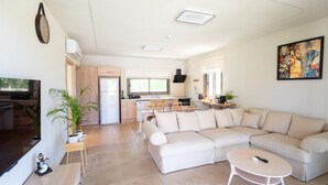 Una sala de estar moderna con un sofá seccional color beige, una mesita de centro y un televisor sobre un mueble de madera. Se ve una cocina con comedor al fondo.