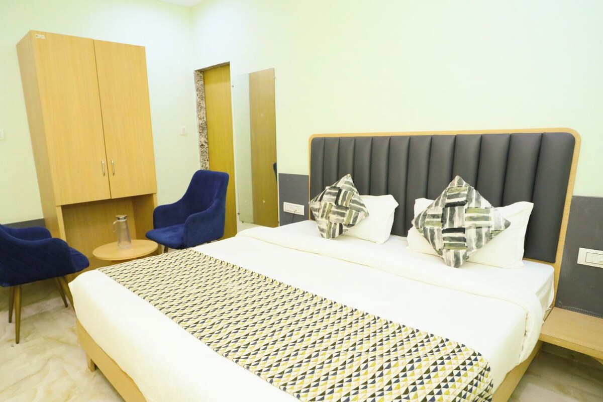 A hotel room with a bed, two blue chairs, a wooden wardrobe, and a small table.