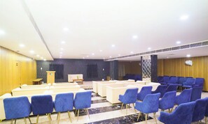A banquet hall with rows of blue chairs, a stage with a podium, and a seating area with sofas. - Hotel Shree Khodiyar By Rangoli Udaipur (Udaipur)