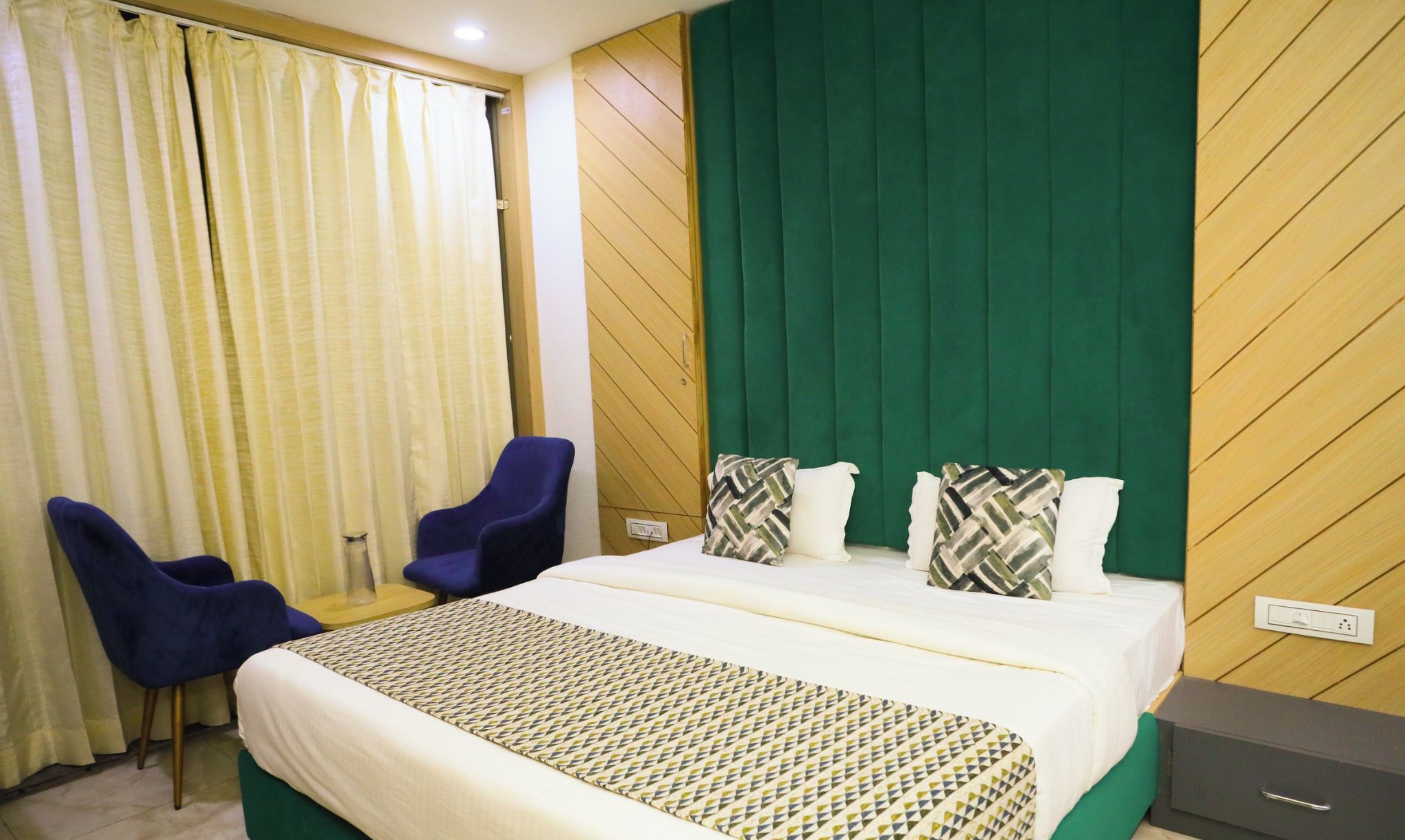 Una habitación de hotel con una cama, dos sillas azules, un cabecero verde y una colcha estampada.