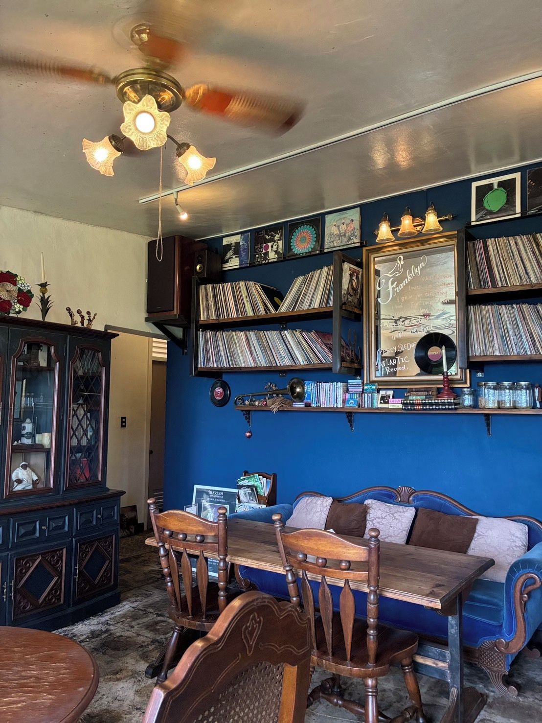 Une pièce avec un mur bleu, un ventilateur de plafond, une table en bois, des chaises, un canapé et une bibliothèque remplie de disques.