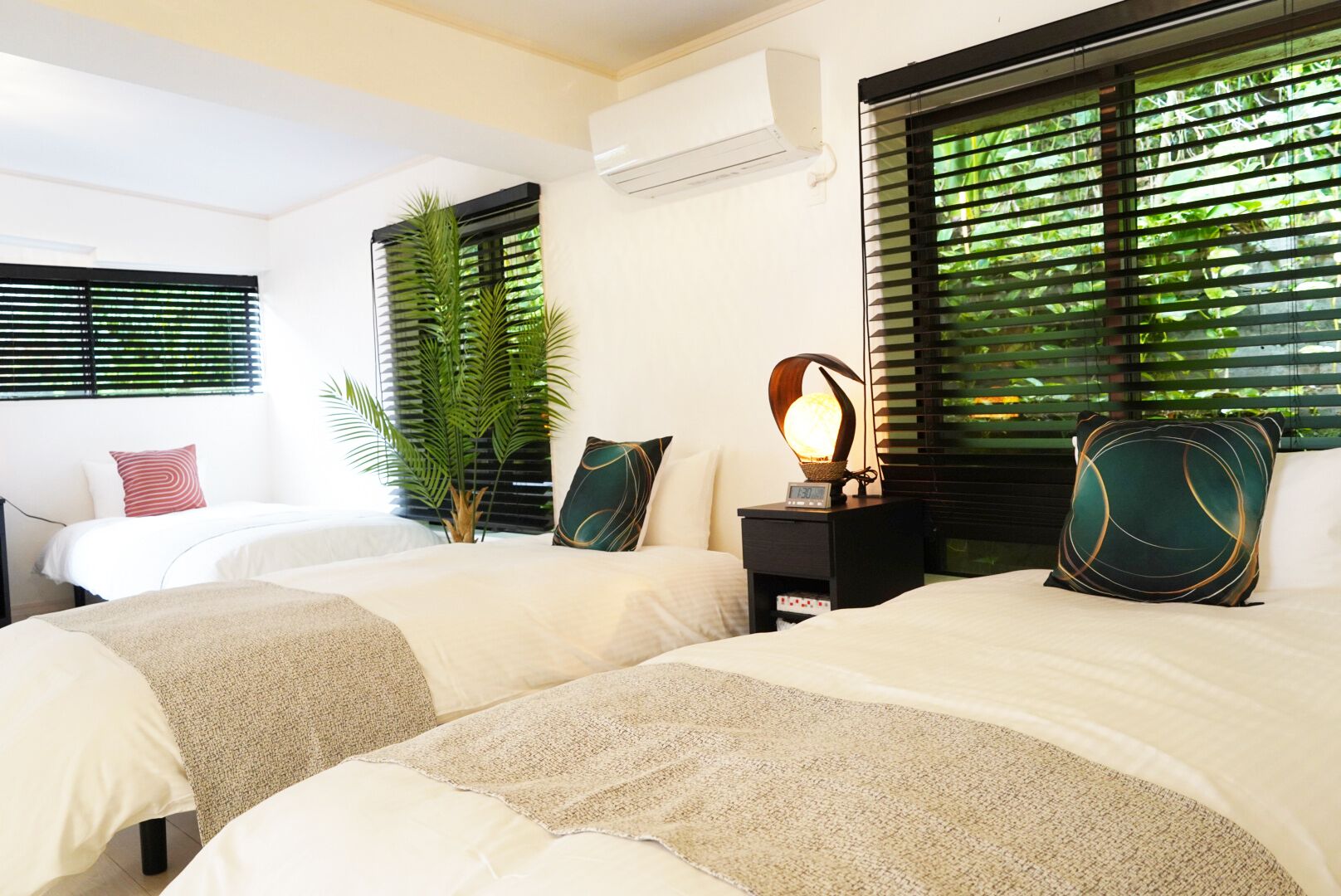 A hotel room with two beds, a nightstand with a lamp, a potted plant, and a window with blinds.