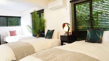 A hotel room with two beds, a nightstand with a lamp, a potted plant, and a window with blinds.