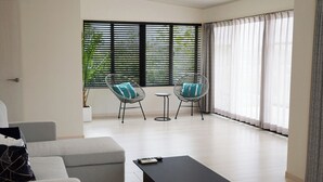 A modern living room with a grey sofa, two armchairs, a coffee table, and a small side table. There are large windows with blinds and sheer curtains, and a potted plant is visible.