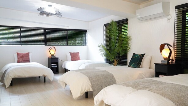 A hotel room with three beds, a large window with blinds, a potted plant, and a wall-mounted air conditioner.