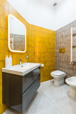 Un baño moderno con pared de azulejos amarillos, un lavamanos blanco con espejo y un inodoro.