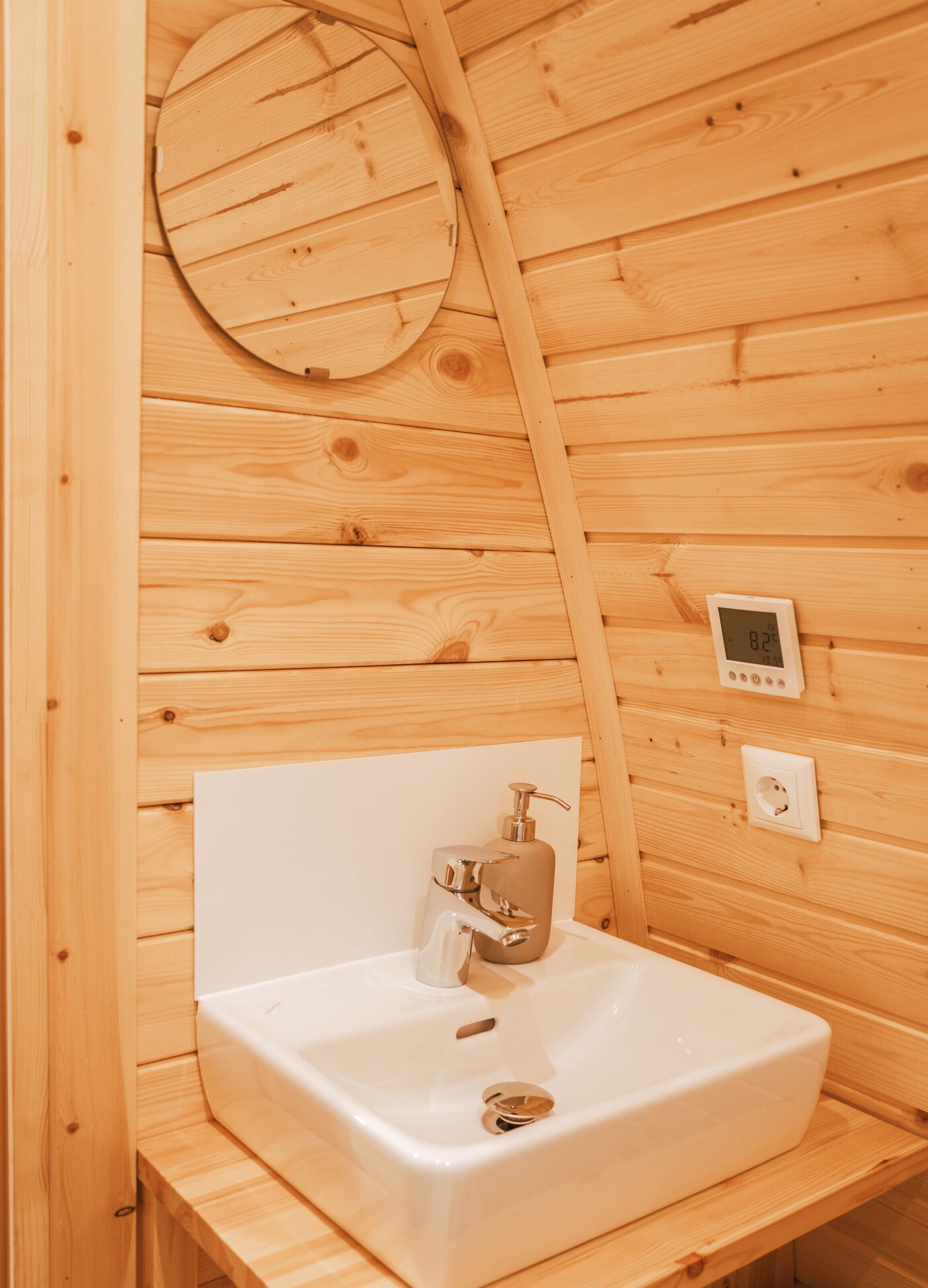 A wooden cabin bathroom with a white sink, a soap dispenser, and a thermostat on the wall.