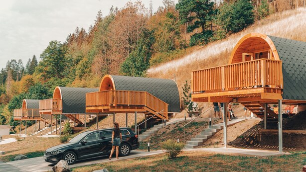 Une rangée de cabanes modernes, semi-ouvertes, dotées de balcons en bois, se dressant sur un fond de feuillage automnal et d’une colline.