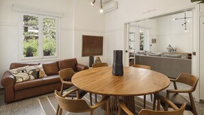 A dining area with a round wooden table, six wooden chairs, a brown leather sofa, and a large window with a view of greenery. - Evas Apartment in the Heart of Chania City (Chania)