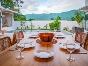 A dining table set for six with a large wooden bowl centerpiece, surrounded by plates and wine glasses, overlooking a pool and lush greenery.