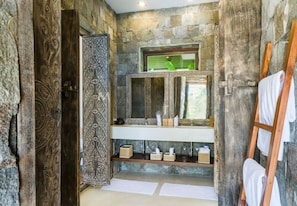 A bathroom with a stone wall, a wooden door with intricate carvings, a mirror above a sink, and a wooden ladder with towels. - FORMOSA-2BR-CHAWENGNOI (Ko Samui)