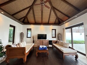 A spacious room with a high ceiling, a ceiling fan, a large flat-screen TV, wooden furniture, and large windows offering a view of the outdoors. - FORMOSA-2BR-CHAWENGNOI (Ko Samui)