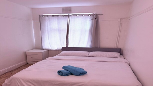A neatly made bed with a white comforter and two folded blue towels. There is a white bedside table with drawers and a white power outlet on the floor.