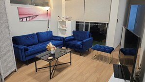 A living room with a blue sofa, a coffee table with a vase, a bookshelf, and a large window. - Stylish 2-Bedroom Apartment in Maddington, 15 Minutes from Perth Airport (Maddington)