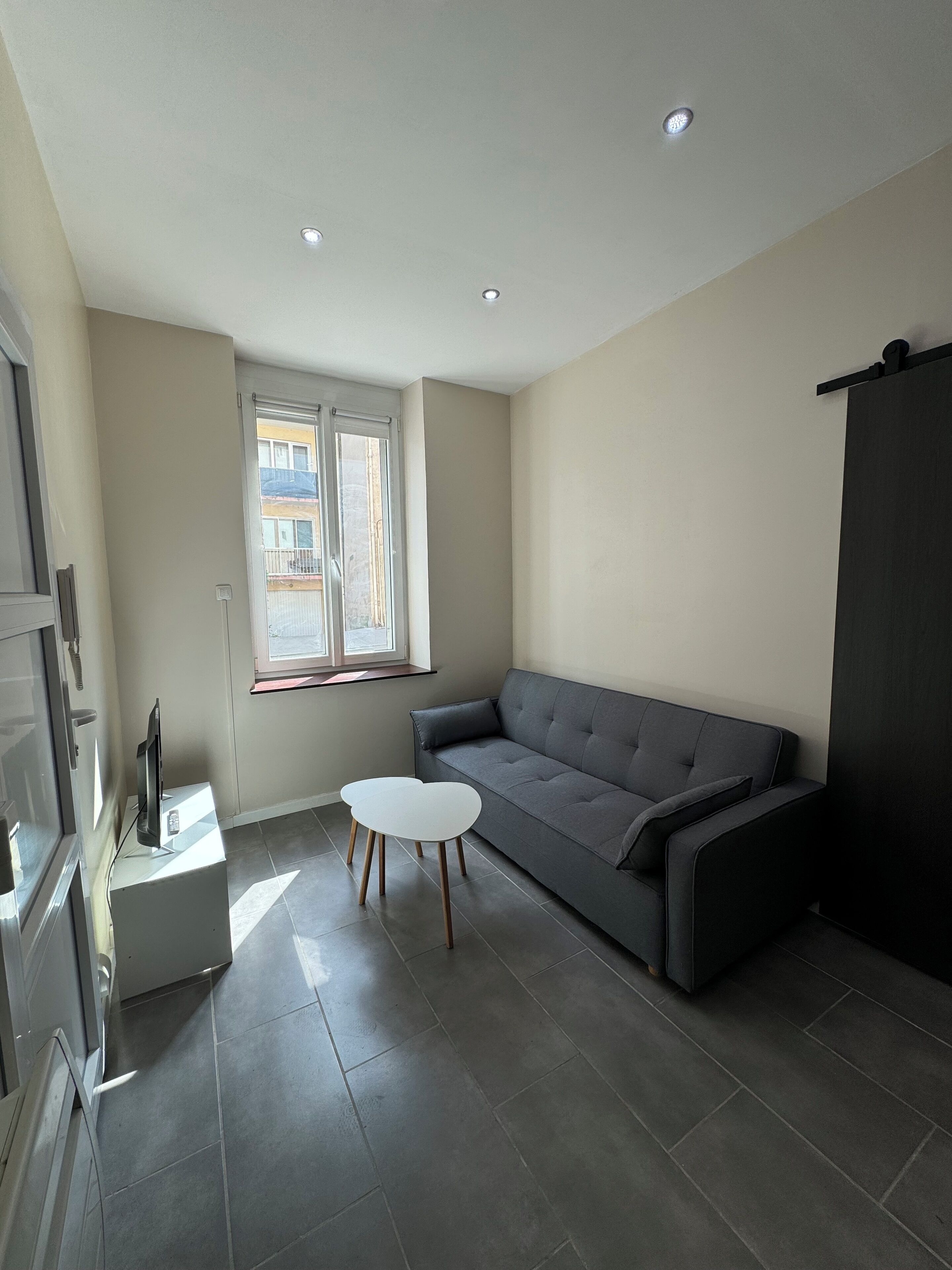 Un salon moderne avec un canapé gris, une petite table blanche et une fenêtre donnant sur des immeubles.