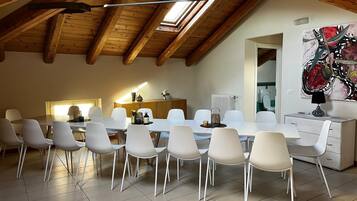 Een eetgedeelte met een hoge plafond en dakraam, witte stoelen, een lange tafel en een moderne keuken op de achtergrond.