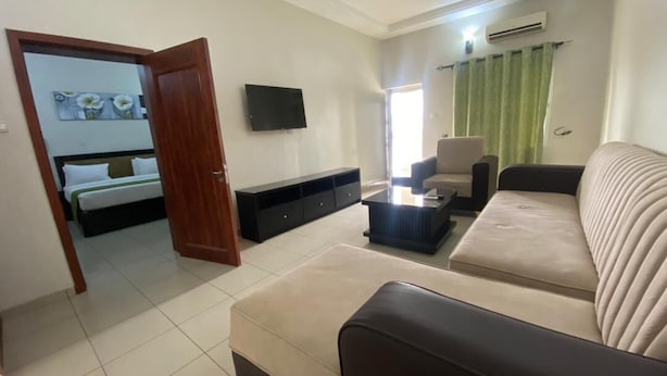 A living room with a sofa, a coffee table, a TV, and a bed in the background. - Stanzel Grand Resort (Abuja)