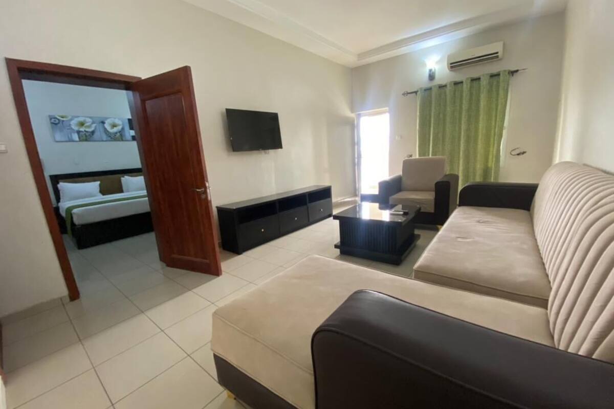 A living room with a sofa, a coffee table, a TV, and a bed in the background.
