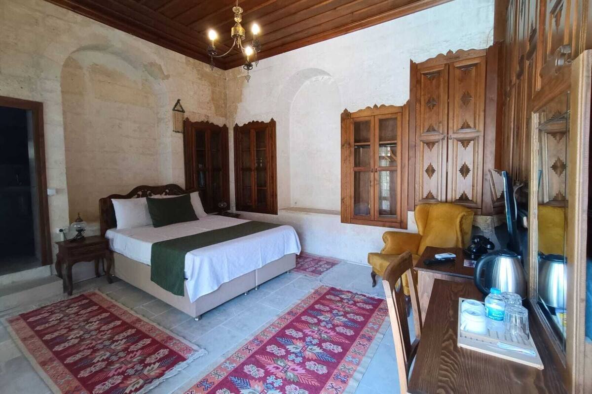 A bedroom with a bed, wooden wardrobe, a chair, a table with a kettle, and a chandelier.