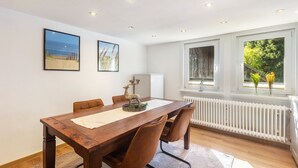 A dining room with a wooden table, chairs, framed pictures, and a window with a view of greenery. - Apartment 'Bödefeld' with Shared Terrace, Private Garden and Wi-Fi (Schmallenberg)