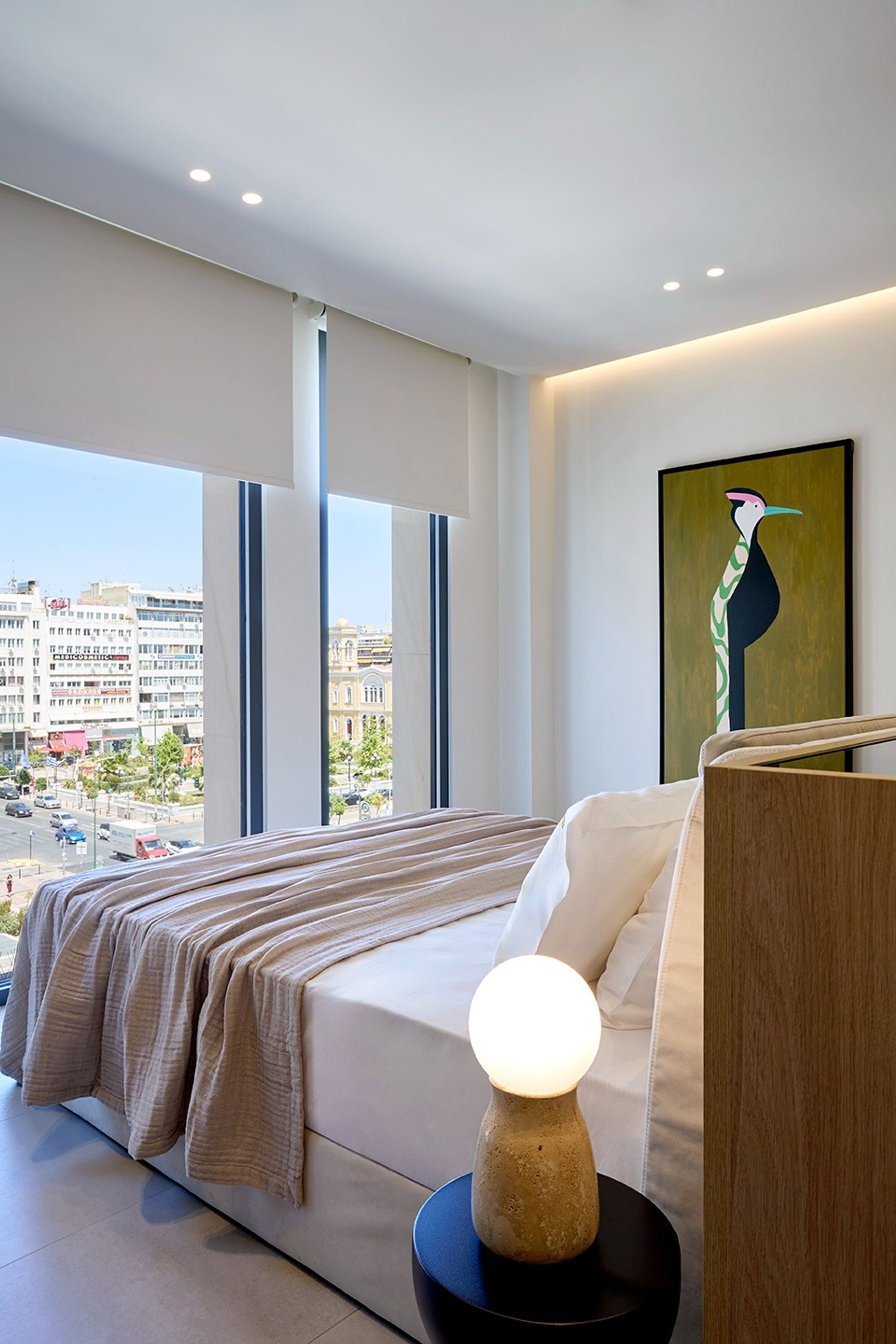 Una habitación de hotel moderna con una cama grande, una mesita de noche, una lámpara y vistas a la ciudad a través de amplias ventanas.