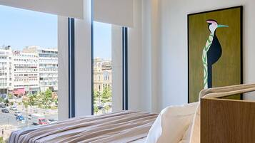 Una habitación de hotel moderna con una cama grande, una mesita de noche, una lámpara y vistas a la ciudad a través de amplias ventanas.